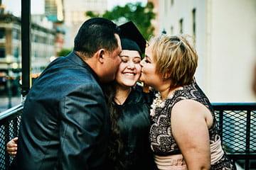 young woman at graduation smiling with her parents kissing her cheeks